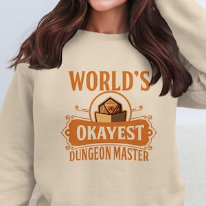 May include: A beige sweatshirt with the text "World's Okayest Dungeon Master" in orange. A graphic of a 20-sided die in a box is above the text.