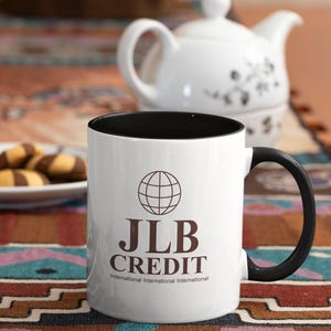 Könnte beinhalten: Weiße Keramiktasse mit schwarzem Rand und Henkel. Die Tasse hat ein braunes Globus-Symbol und den Text "JLB CREDIT International International International" aufgedruckt.