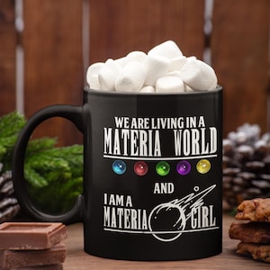 Może przedstawiać: Czarny ceramiczny kubek wypełniony białymi piankami. Na kubku widnieje biały napis "WE ARE LIVING IN A MATERIA WORLD AND I AM A MATERIA GIRL". Kubek stoi na drewnianej powierzchni z czekoladą i ciastkami.