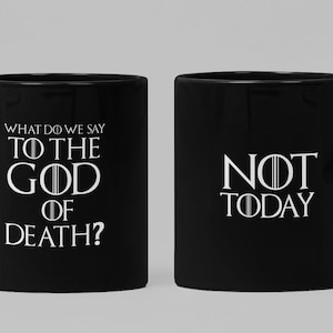 Pode incluir: Duas canecas de café pretas com texto branco. Uma caneca diz "O que dizemos ao deus da morte?" e a outra caneca diz "Não hoje".