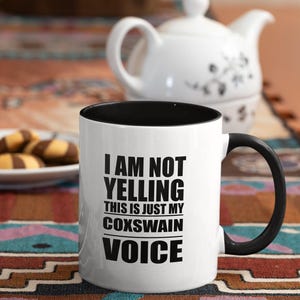 May include: White coffee mug with a black rim and handle. The mug has black text that reads "I am not yelling this is just my coxswain voice".