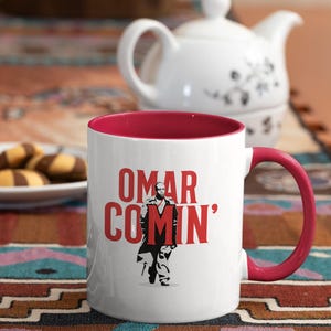 May include: White ceramic mug with a red rim and handle. The mug features a black and white illustration of a man holding a red letter "M" with the text "OMAR COMIN'" in red letters.