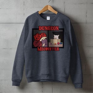 May include: A dark gray sweatshirt with a white cat and a person with dice for a head. The text on the sweatshirt says "Dungeon Meowster".