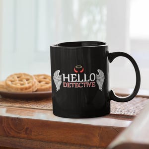Pode incluir: Caneca de cerâmica preta com um design de asas de anjo brancas e o texto "HELLO DETECTIVE" em branco. A caneca tem uma auréola vermelha acima do texto.