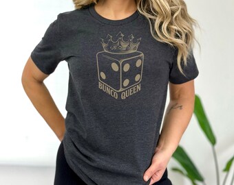 Op de afbeelding: Donkergrijs t-shirt met een goudkleurige afbeelding van een dobbelsteen, een kroon en de tekst "Bunco Queen". Het shirt heeft een ronde hals en is gemaakt van zacht materiaal.