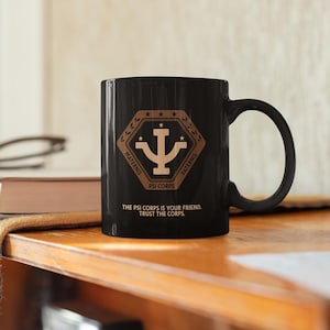 Op de afbeelding: Zwarte keramische mok met een bruine afbeelding van een gestileerd symbool met de tekst "PSI CORPS" en de woorden "MATERNIS" en "PATERNIS" aan weerszijden. De tekst "THE PSI CORPS IS YOUR FRIEND. TRUST THE CORPS." staat onder het symbool gedrukt.