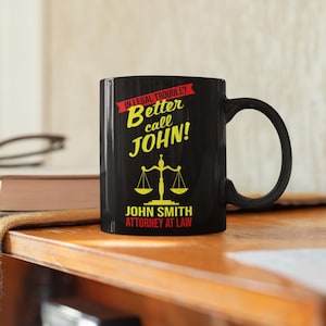 May include: Black coffee mug with a yellow and red design. The mug says "In legal trouble? Better call John!" and "John Smith Attorney at Law".