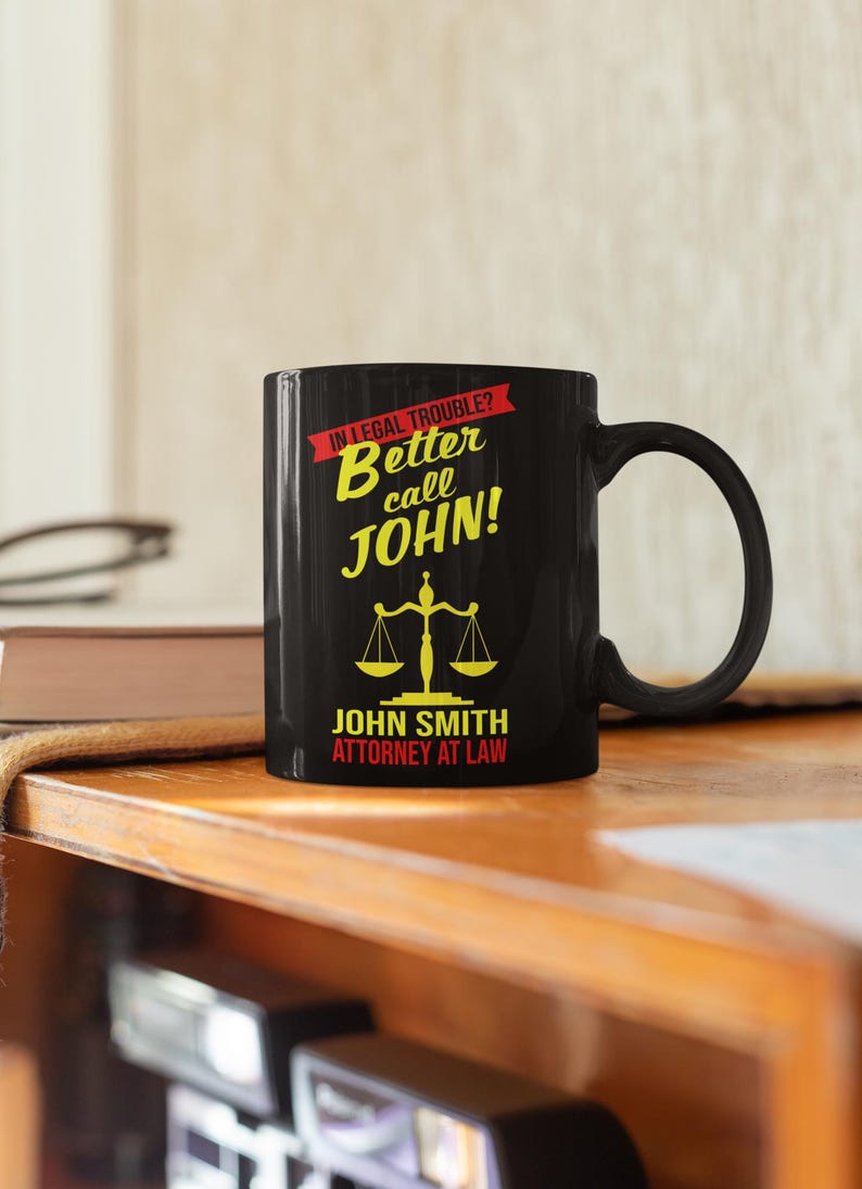 May include: Black coffee mug with a yellow and red design. The mug says "In legal trouble? Better call John!" and "John Smith Attorney at Law".