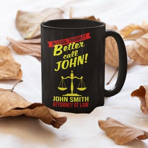 May include: Black coffee mug with a white background. The mug has a graphic that says "In legal trouble? Better call JOHN!" with a scale of justice and the text "JOHN SMITH ATTORNEY AT LAW".