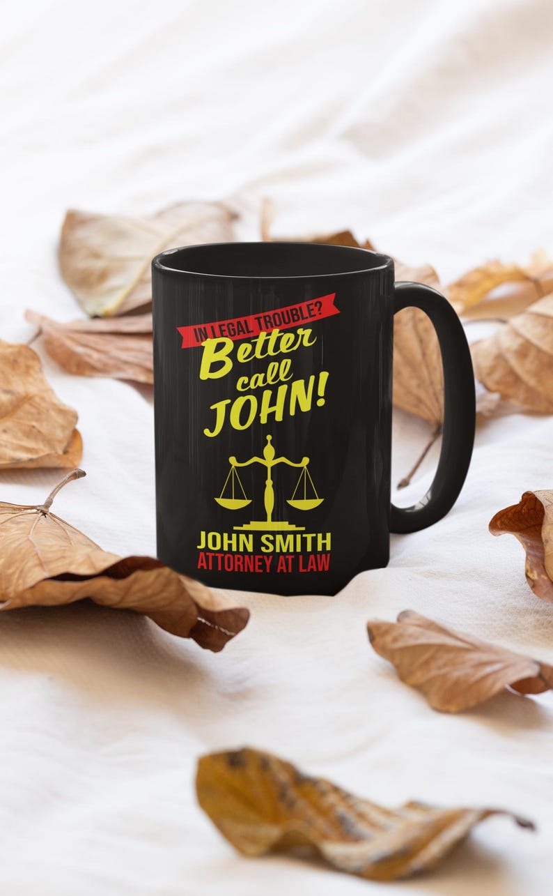 May include: Black coffee mug with a white background. The mug has a graphic that says "In legal trouble? Better call JOHN!" with a scale of justice and the text "JOHN SMITH ATTORNEY AT LAW".