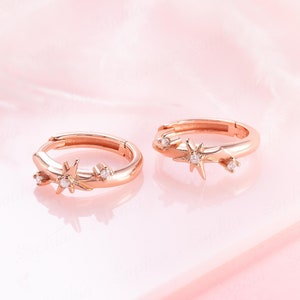 May include: A pair of rose gold hoop earrings. Each earring has a starburst design with small, clear gemstones. The earrings are set against a soft pink background, creating a delicate and elegant aesthetic.