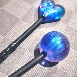 May include: Two handcrafted wands with celestial-themed designs. One wand features a heart-shaped top, the other a sphere. Both have dark, textured handles and are filled with swirling blue and purple colors, creating a galaxy effect.
