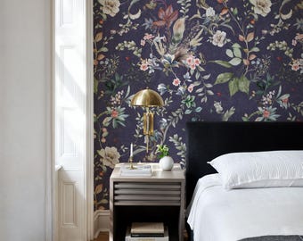May include: A bedroom with a white nightstand and a bed with a white duvet and a black throw blanket. The wall behind the bed is covered in a dark purple floral wallpaper with white flowers and green leaves.
