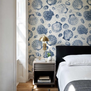 May include: A bedroom with a white and blue seashell wallpaper, a black headboard bed with white bedding, a nightstand with a gold lamp, and a white vase with a plant.