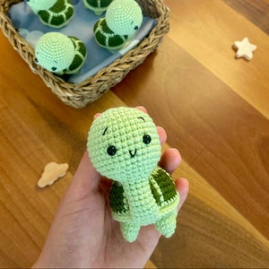 May include: A light green crocheted turtle with a dark green shell pattern and black eyes. Several similar turtles are in a wicker basket. The turtle is held in a hand. A wooden star and cloud are in the background.