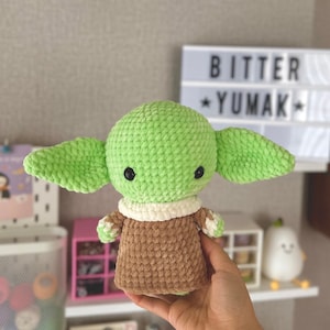 May include: A crocheted doll of a character with large ears, green skin, and black eyes. The doll wears a brown robe with a white collar. The text "BITTER YUMAK" is visible in the background.