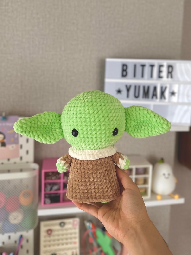 May include: A crocheted doll of a character with large ears, green skin, and black eyes. The doll wears a brown robe with a white collar. The text "BITTER YUMAK" is visible in the background.
