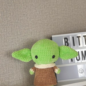 May include: A handmade, green and brown crocheted doll with large ears and black eyes. The doll is wearing a cream-colored collar and a brown robe. The doll is displayed on a white shelf against a gray wall.
