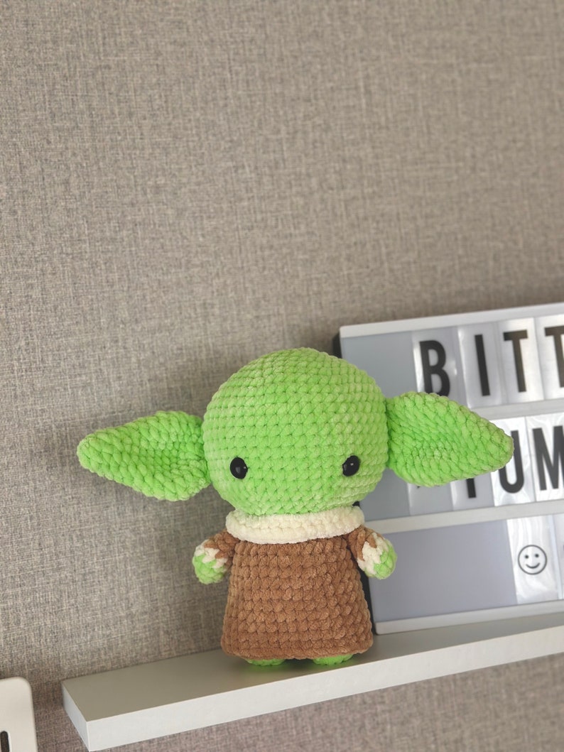 May include: A handmade, green and brown crocheted doll with large ears and black eyes. The doll is wearing a cream-colored collar and a brown robe. The doll is displayed on a white shelf against a gray wall.