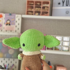 May include: A handmade green crocheted doll with large ears, black eyes, and a brown and white outfit. The doll is a character from a popular science fiction franchise. The doll is approximately 6 inches tall.