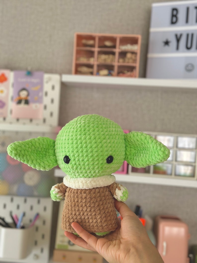 May include: A handmade green crocheted doll with large ears, black eyes, and a brown and white outfit. The doll is a character from a popular science fiction franchise. The doll is approximately 6 inches tall.
