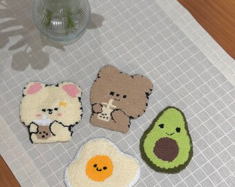 May include: Four handmade, tufted coasters in the shape of a bear, avocado, and fried egg. The bear coasters are beige and brown, the avocado is green, and the egg is yellow and white. The coasters are placed on a white grid patterned surface.