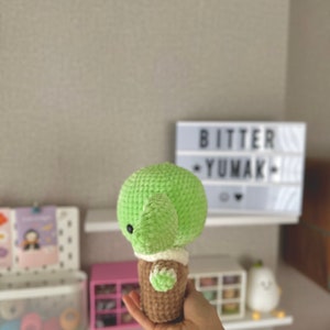 May include: A handmade, green crocheted turtle with a brown shell and white accents. The turtle is held in a hand. The text "BITTER YUMAK" is visible in the background on a light box.