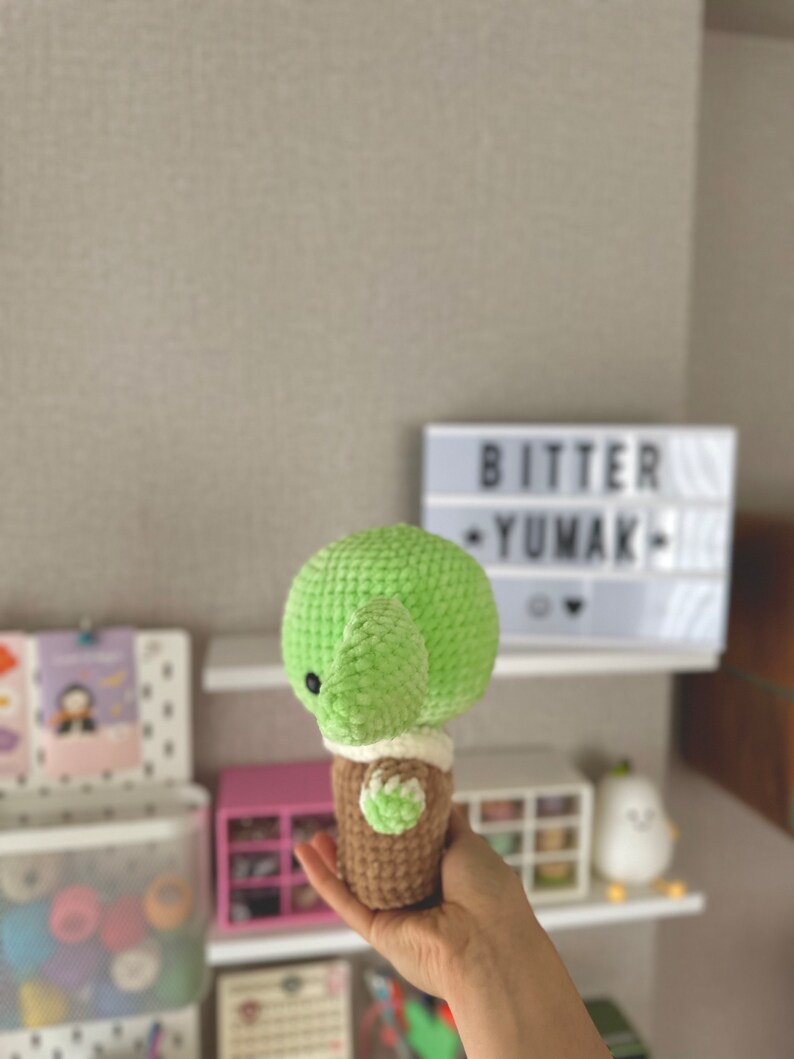 May include: A handmade, green crocheted turtle with a brown shell and white accents. The turtle is held in a hand. The text "BITTER YUMAK" is visible in the background on a light box.