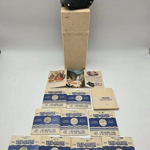 May include: A vintage View-Master set, featuring a black viewer, a beige storage case, and multiple reels. The reels are in blue and white packaging, with text that reads "7 More Wonders of the World".