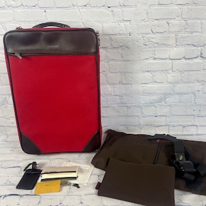 May include: A red Hartmann luggage suitcase with brown leather accents and a black luggage tag. The suitcase is sitting on a white brick wall background. There is a brown garment bag and a black luggage strap in the foreground.