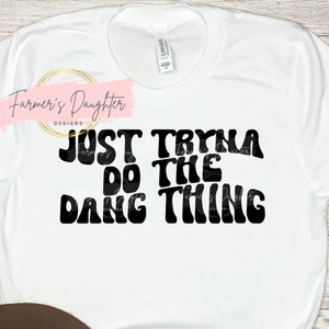 May include: White t-shirt with black text that reads "Just Tryna Do The Dang Thing" in a retro font style.