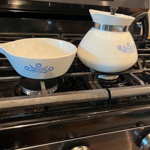 May include: A white ceramic teapot with a black handle and lid, featuring a blue floral pattern, alongside a matching white ceramic bowl with a handle and the same blue floral design, both resting on a black metal stovetop grate.