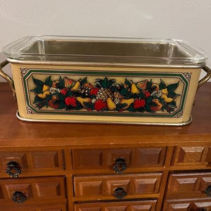 May include: A vintage yellow metal baking dish with a glass lid. The dish features a colorful fruit design with apples, pears, pineapples, and other fruits.