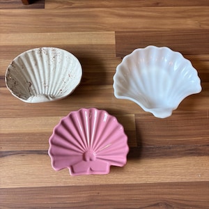 May include: Three shell-shaped dishes arranged together. One dish is white with a brown marbled pattern, one is solid white, and one is solid pink. Each dish features a scalloped edge and a ribbed texture radiating from the center hinge area.