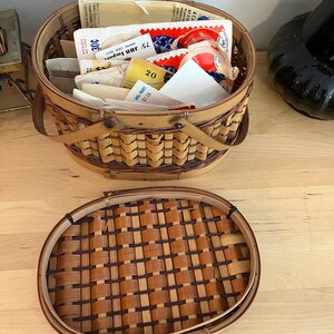 May include: A woven basket with a hinged lid, filled with various papers and small items. The basket is made of light and dark brown woven material. The lid is open, revealing the contents, including papers with text and numbers. The basket is on a light wood surface.