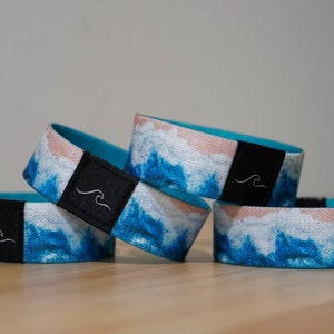 May include: Four blue and white patterned wristbands with a black band and a white wave logo. The wristbands are stacked on top of each other.