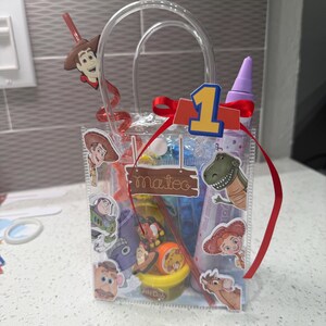 K&ouml;nnte beinhalten: Eine transparente Geschenk-Tasche mit Henkel, verziert mit Toy Story-Charakter-Aufklebern und einem roten Band. Die Tasche enth&auml;lt Spielzeug und einen Strohhalm mit einer Woody-Figur. Eine 1 und der Name "Mateo" sind ebenfalls sichtbar.