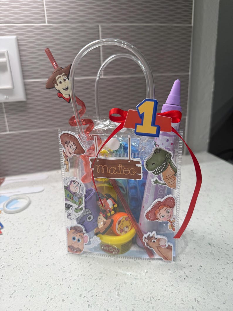 K&ouml;nnte beinhalten: Eine transparente Geschenk-Tasche mit Henkel, verziert mit Toy Story-Charakter-Aufklebern und einem roten Band. Die Tasche enth&auml;lt Spielzeug und einen Strohhalm mit einer Woody-Figur. Eine 1 und der Name "Mateo" sind ebenfalls sichtbar.