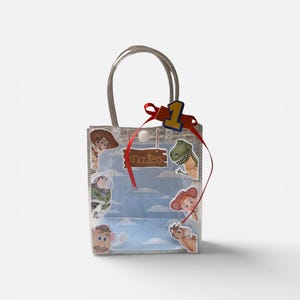 K&ouml;nnte beinhalten: Transparente Geschenkt&uuml;te mit silbernen Griffen, verziert mit Toy Story-Charakteraufklebern. Ein rotes Band und die Zahl eins sind oben angebracht. Der Name "Mateo" steht auf einem Holzschild.