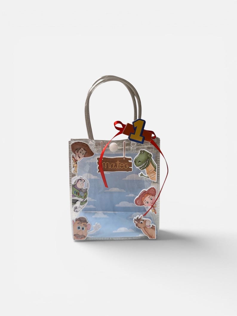 K&ouml;nnte beinhalten: Transparente Geschenkt&uuml;te mit silbernen Griffen, verziert mit Toy Story-Charakteraufklebern. Ein rotes Band und die Zahl eins sind oben angebracht. Der Name "Mateo" steht auf einem Holzschild.