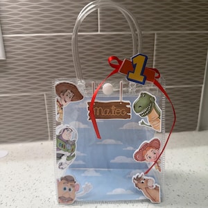 K&ouml;nnte beinhalten: Transparente Plastik-Geschenkt&uuml;te mit Toy Story-Figuren, darunter Woody, Buzz Lightyear und Rex. Die T&uuml;te hat ein rotes Band, die Zahl eins und den Namen "Mateo". Die T&uuml;te hat einen transparenten Griff.