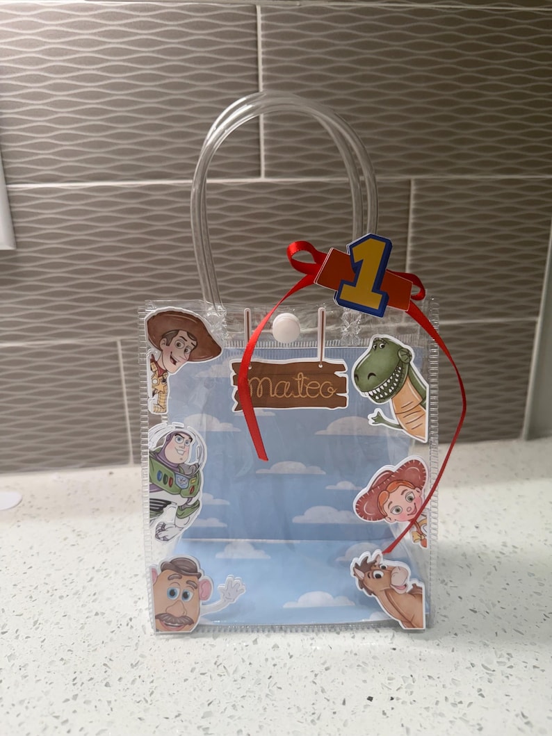 K&ouml;nnte beinhalten: Transparente Plastik-Geschenkt&uuml;te mit Toy Story-Figuren, darunter Woody, Buzz Lightyear und Rex. Die T&uuml;te hat ein rotes Band, die Zahl eins und den Namen "Mateo". Die T&uuml;te hat einen transparenten Griff.