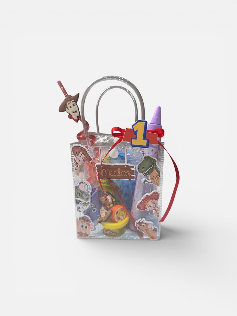 K&ouml;nnte beinhalten: Eine transparente Plastik-Geschenkt&uuml;te gef&uuml;llt mit Partyzubeh&ouml;r zum Thema Toy Story. Die Tasche hat ein rotes Band, eine Eins und einen Woody-f&ouml;rmigen Lolli. Aufkleber von Charakteren wie Buzz Lightyear und Rex sind sichtbar.