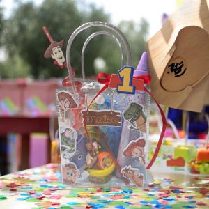 K&ouml;nnte beinhalten: Transparente Party-Geschenkt&uuml;te mit Aufklebern von Toy Story-Charakteren. Die Tasche enth&auml;lt Spielzeug und einen Play-Doh-Beh&auml;lter. Ein rotes Band und eine Eins-Aussparung schm&uuml;cken die Oberseite. Der Name "Mateo" steht auf einem Holzschild.