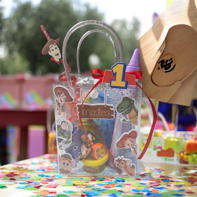 K&ouml;nnte beinhalten: Transparente Party-Geschenkt&uuml;te mit Aufklebern von Toy Story-Charakteren. Die Tasche enth&auml;lt Spielzeug und einen Play-Doh-Beh&auml;lter. Ein rotes Band und eine Eins-Aussparung schm&uuml;cken die Oberseite. Der Name "Mateo" steht auf einem Holzschild.