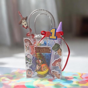 K&ouml;nnte beinhalten: Transparente Plastik-Geschenkt&uuml;te, verziert mit Toy Story-Charakteraufklebern und einem roten Band. Die Tasche hat einen transparenten Griff und enth&auml;lt verschiedene Spielsachen und Leckereien. Eine 1 und der Name "Mateo" sind ebenfalls sichtbar.