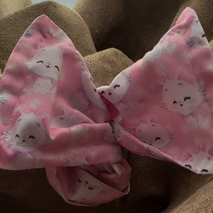 May include: A pink fabric bow with a white cat print. The bow is tied in a knot and has a ruffled edge.