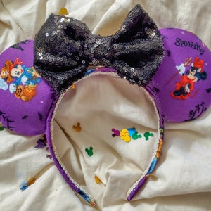 May include: Purple Minnie Mouse ears with a black sequined bow. The ears feature a Halloween theme with Mickey Mouse and Minnie Mouse characters. The text "Spooky" is printed on one ear.