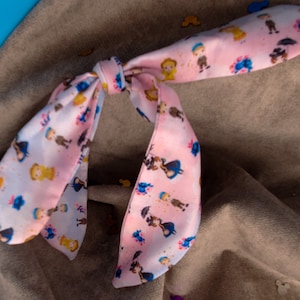 Puede incluir: Una diadema de tela rosa con un lazo, que presenta un patrón repetido de pequeñas figuras de dibujos animados en azul, amarillo y blanco.