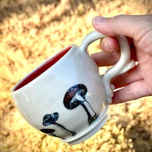 May include: White ceramic mug with a red interior and a unique handle design. The mug features a hand-painted mushroom design in shades of blue, brown, and white. The mug is held in a hand against a blurred background of dry grass.